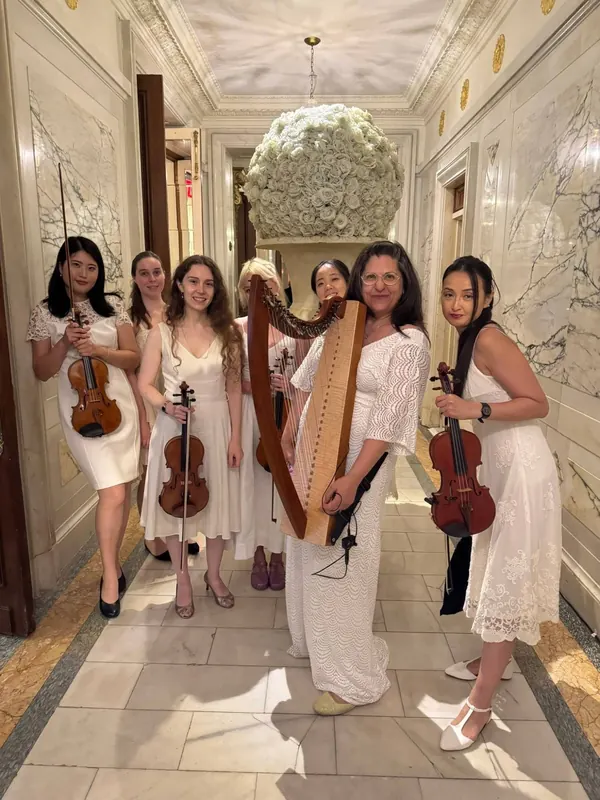 Mia Theodoratus with her lever harp and a string ensemble, all dressed in white, in a marble hallway