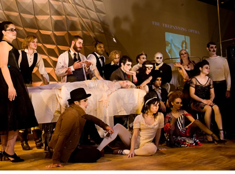 The Citizens Band in costume around a figure on a table, The Trepanning Opera projected behind