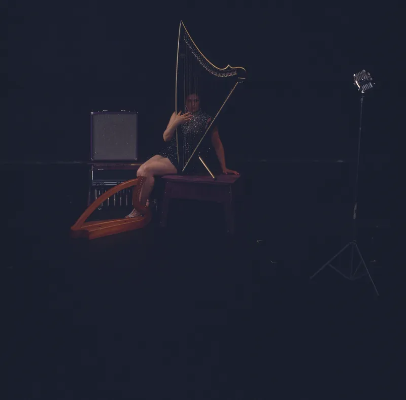 Mia seated on a stage behind an electric harp, a lever harp at her feet and a vintage microphone to the side